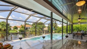 A glass-enclosed outdoor pool area with a table and chairs, a glass of wine, and a view of the surrounding landscape.