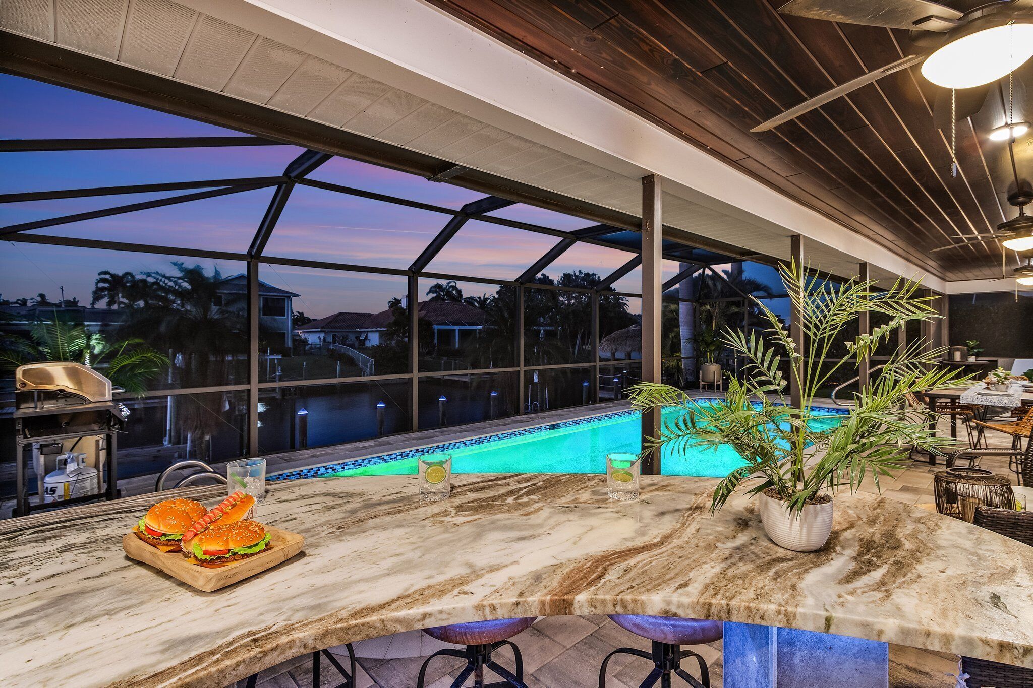 A bar area with a pool view, a grill, and a dining table with burgers.