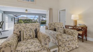 Ein Wohnzimmer mit sofas in floralem Muster, einem Holztisch und Blick auf einen Pool und Palmen durch eine offene Tür.