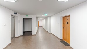 A long, narrow hallway with white walls and a tiled floor, leading to an elevator and a door marked with the number 207.