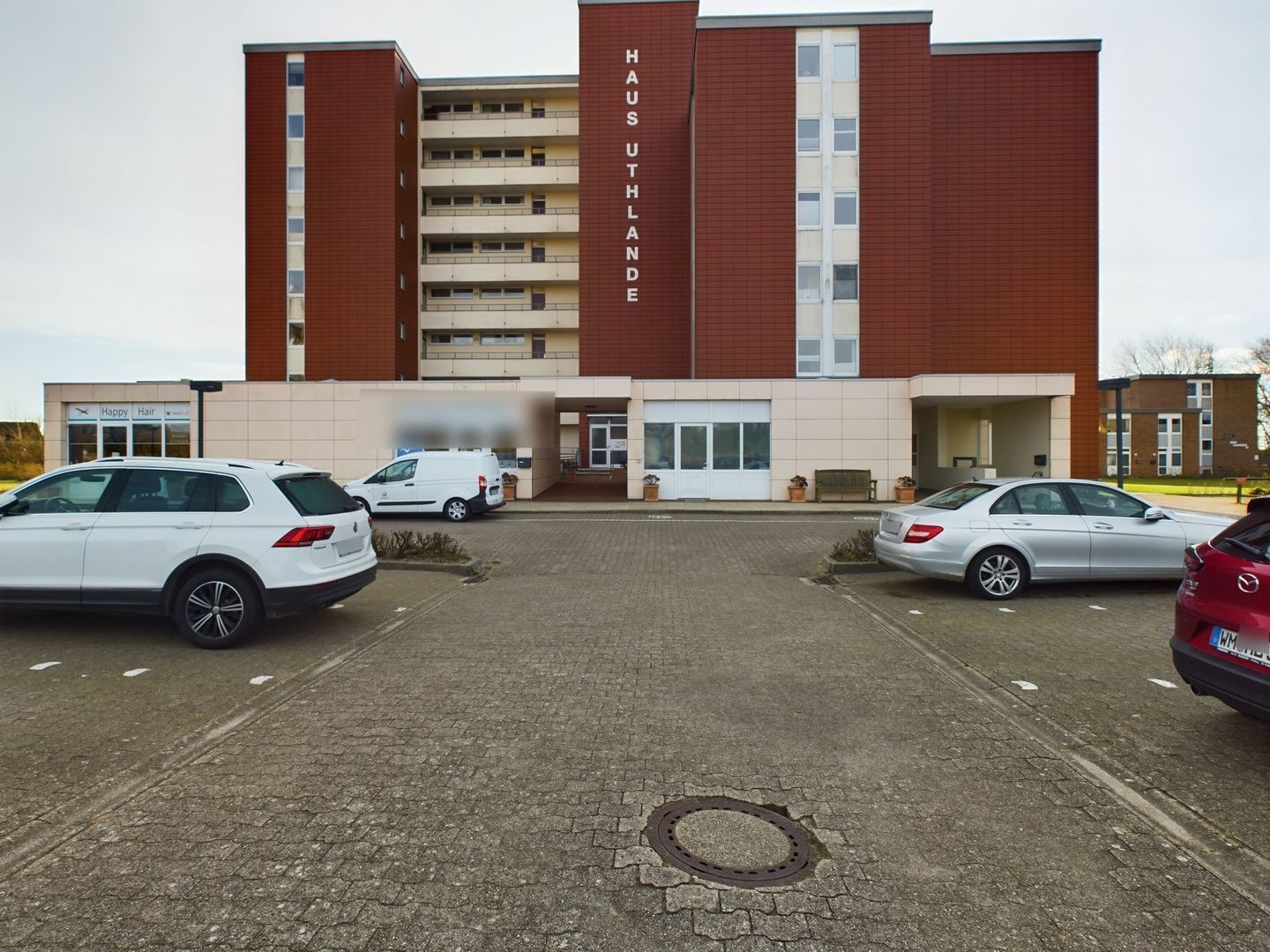 Un hotel de diverses plantes amb façana vermella i beix, un aparcament amb diversos cotxes, i un rètol que diu "Happy Hair".