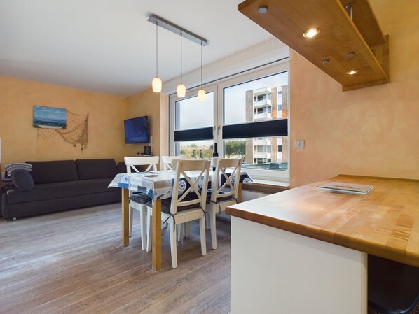 A modern kitchen with a wooden countertop, a dining area with white chairs, a sofa, and a TV mounted on the wall.