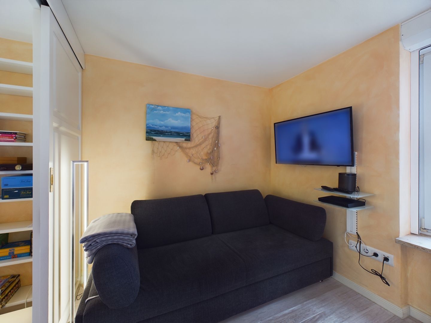 Ein Wohnzimmer mit einem dunklen Sofa, einem an der Wand montierten Fernseher und einem Bücherregal mit Büchern und Dekorationsgegenständen.