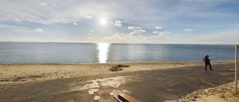 Una playa con una persona caminando, cielo despejado y el sol reflejándose en el agua.