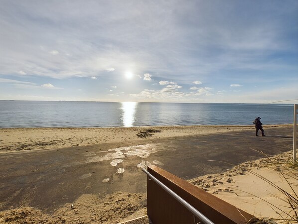 Een strand met een persoon die loopt, een heldere lucht en de zon die op het water reflecteert.