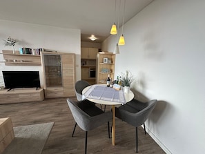 Una sala moderna con una mesa de comedor redonda, dos sillas grises, un televisor, una estantería y una zona de cocina.