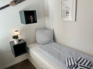 Een compacte, verzorgde slaapkamer met een enkel bed, een nachtkastje met een lamp en een plant, een klein plankje met boeken, en een lijstje met een foto aan de muur.