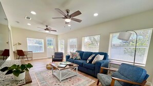 A spacious living room with a blue sofa, a wooden coffee table, and a blue armchair. There are two ceiling fans, a large window with blinds, and a floor lamp. - Pool house 3 bed 2 bath, 1 mile from the beach (Largo)