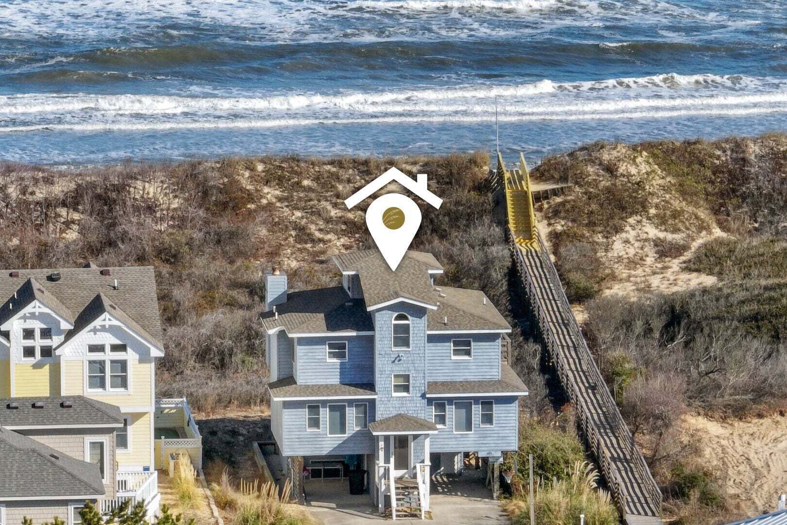 A coastal house with a prominent location marker, a wooden boardwalk leading to the beach, and a view of the ocean.