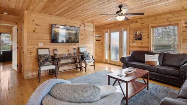 L’intérieur chaleureux d’un chalet avec des murs en bois, un ventilateur de plafond, un grand téléviseur, un canapé en cuir et une table basse en bois.