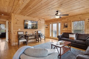 Ein gemütliches Blockhaus-Interieur mit Holzwänden, Deckenventilator, großem Fernseher, Ledersofa und Couchtisch.