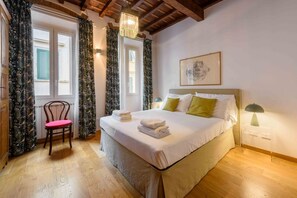 Un dormitorio con una cama, una silla, un ventanal con contraventanas y una lámpara.