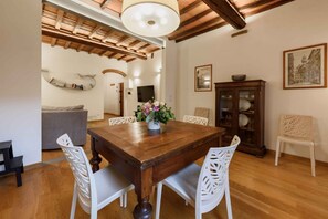 Un comedor con una mesa de madera, sillas blancas, un jarrón con flores, un armario con vajilla y un televisor al fondo.