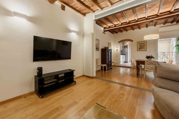 Een woonkamer met een flatscreen-tv, houten vloeren, een bank en een eethoek met stoelen.