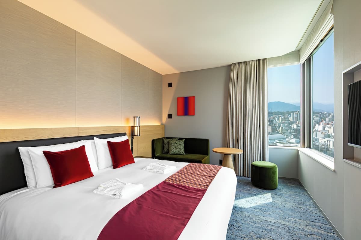 Quarto de hotel com uma cama grande, um sofá verde, uma mesa redonda pequena e vista da cidade através de amplas janelas.