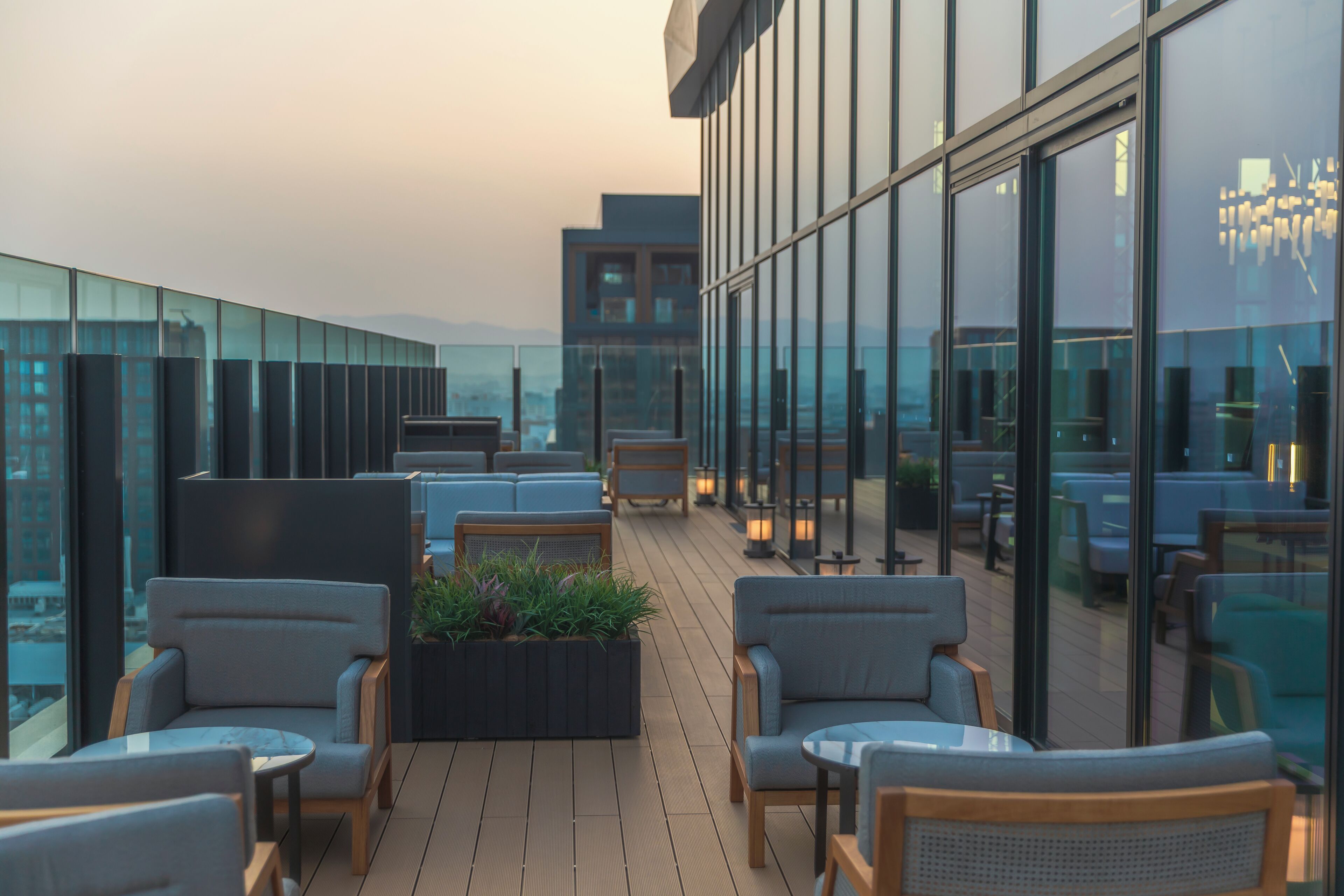 A modern rooftop terrace with seating areas, a planter with plants, and a view of the cityscape through large windows.
