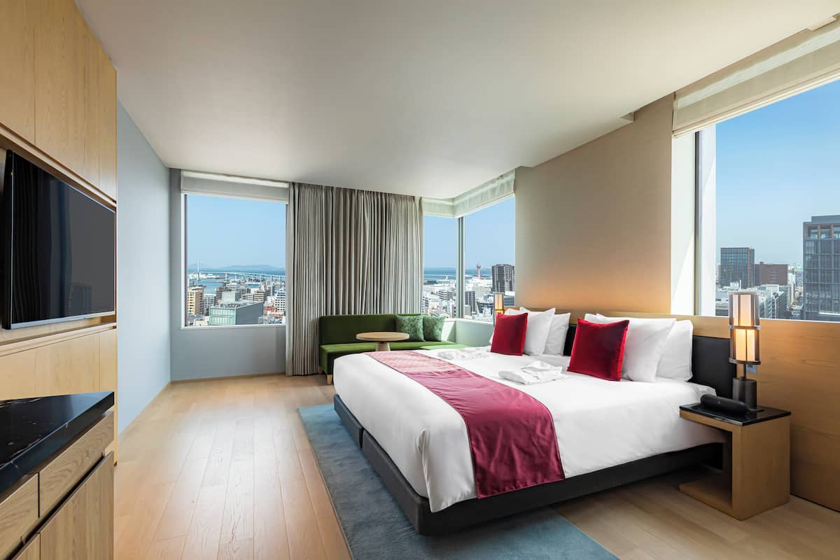 Quarto de hotel moderno com uma cama grande, televisão, vista para a cidade e um sofá verde.
