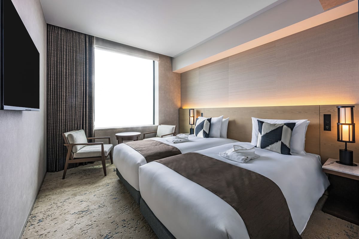 Quarto de hotel com duas camas, uma mesa, uma cadeira, televisão e uma janela grande.