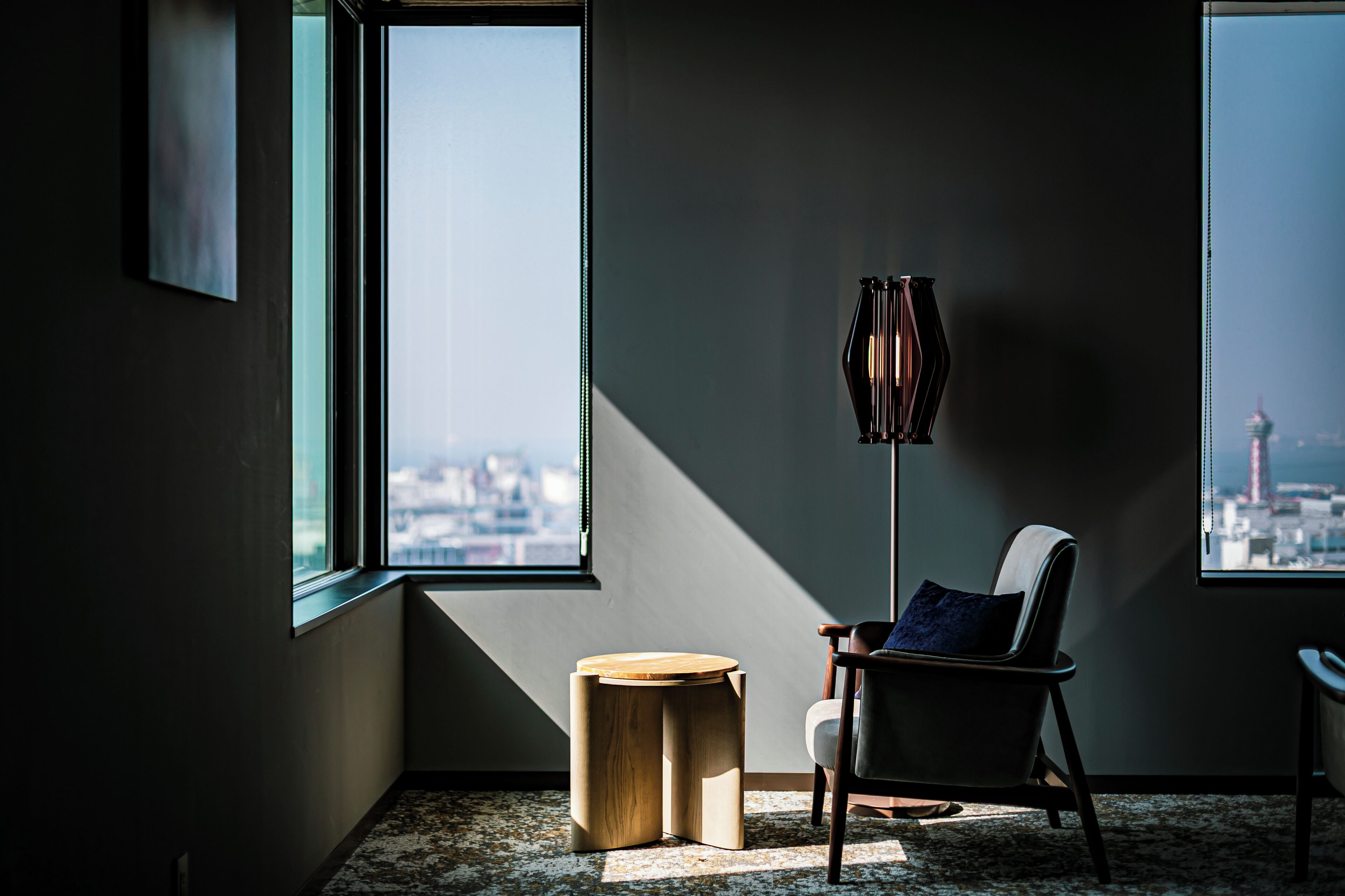 Una habitación moderna con un ventanal grande, una lámpara de pie, un taburete de madera redondo y una silla con cojín.
