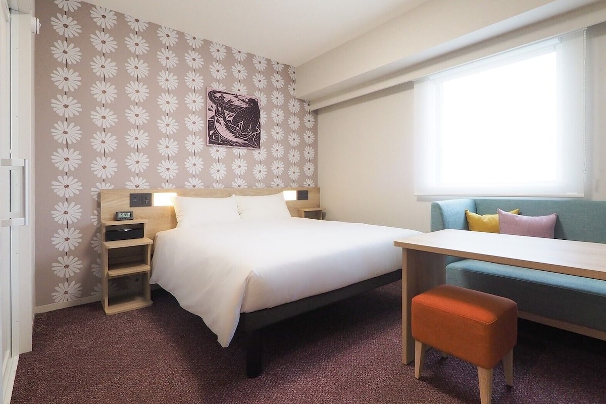 Quarto de hotel com cama, escrivaninha, cadeira, sofá, relógio de parede e janela.