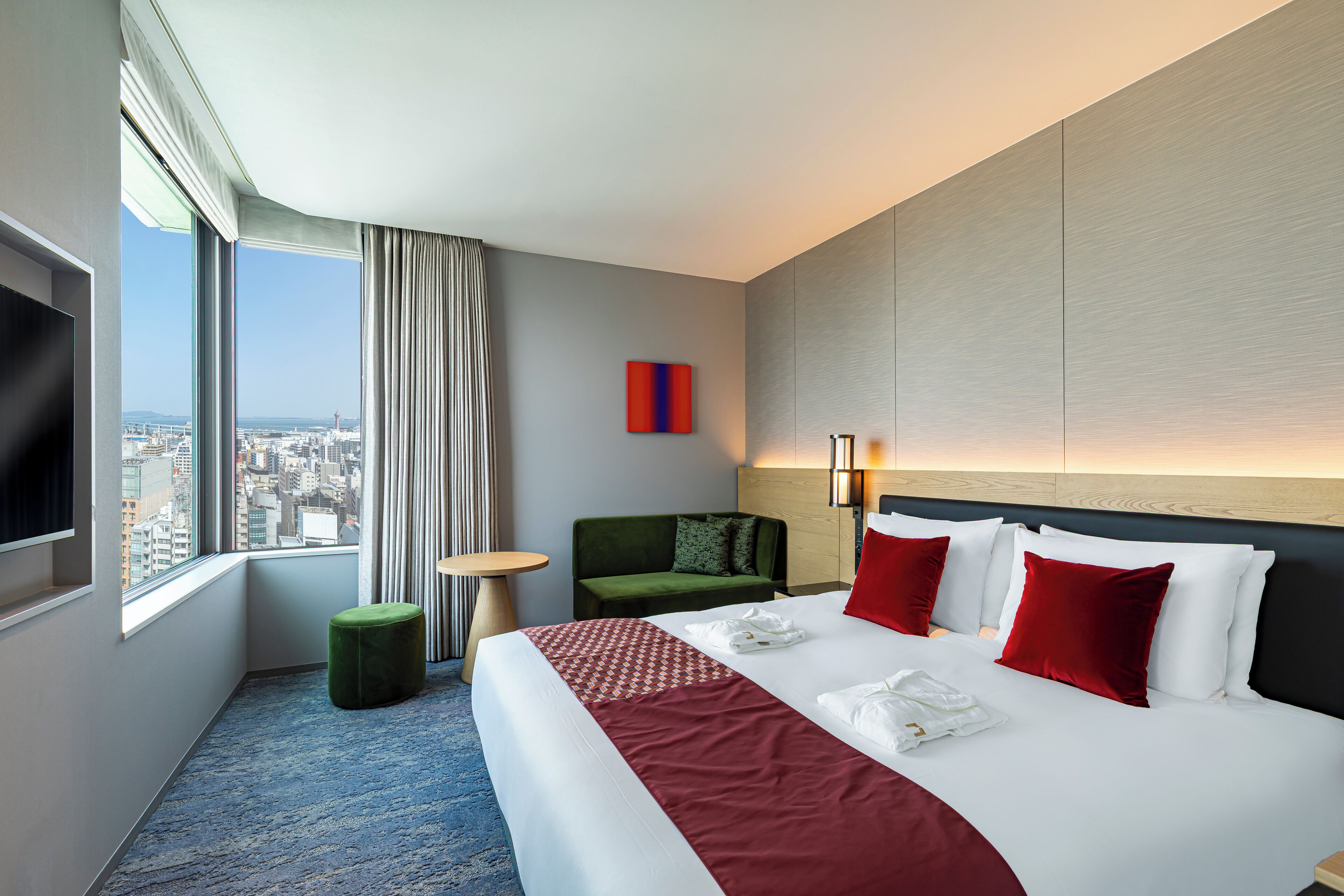 A hotel room with a large bed, a green armchair, a small round table, and a TV on the left wall.