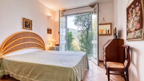 Une chambre à coucher comprenant un lit, une chaise, une table de chevet, une lampe, un miroir et une vue sur les arbres à travers une porte vitrée.