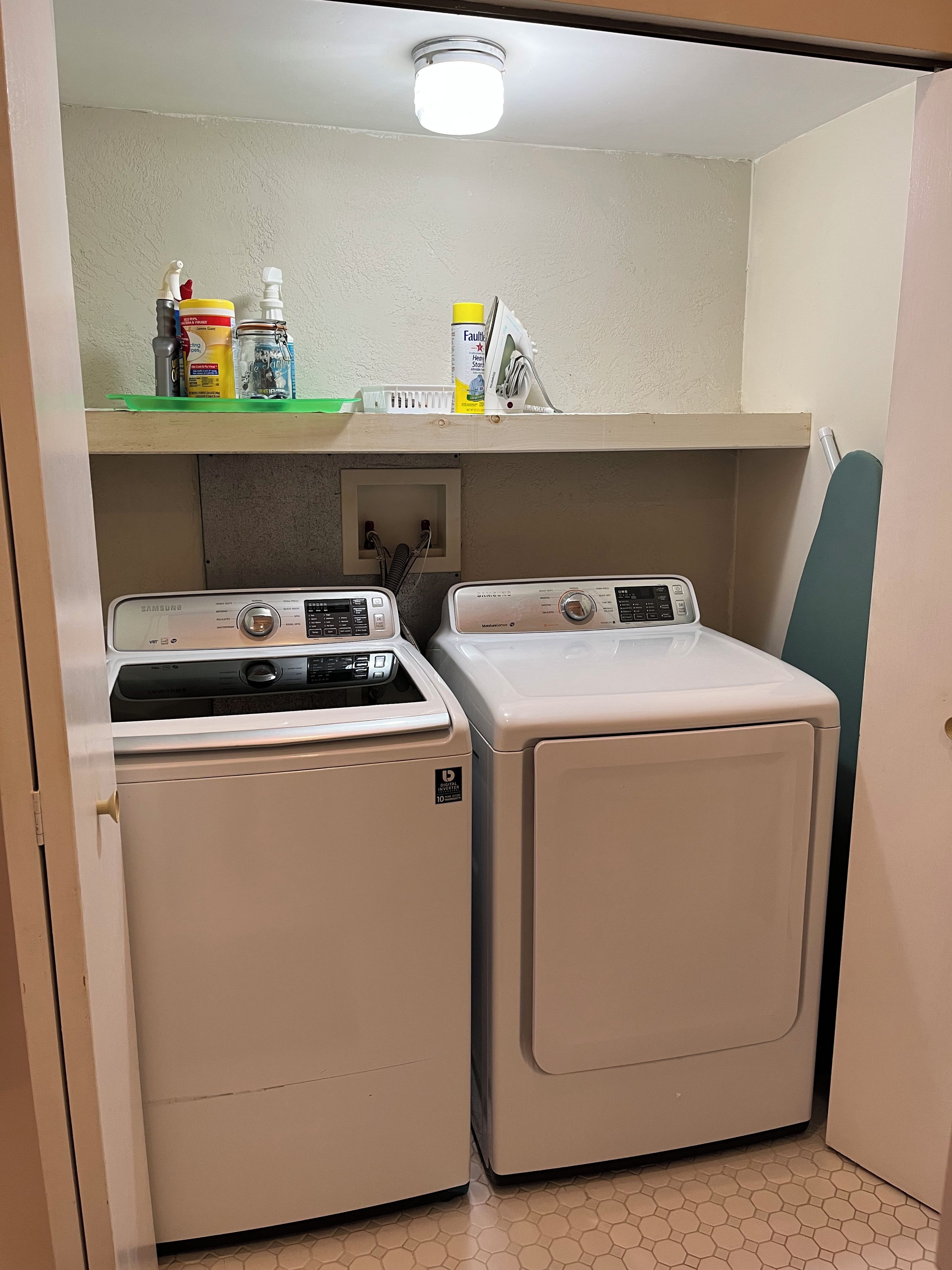 A laundry room with two front-loading washers and dryers, a shelf with cleaning supplies, and a visible electrical outlet.