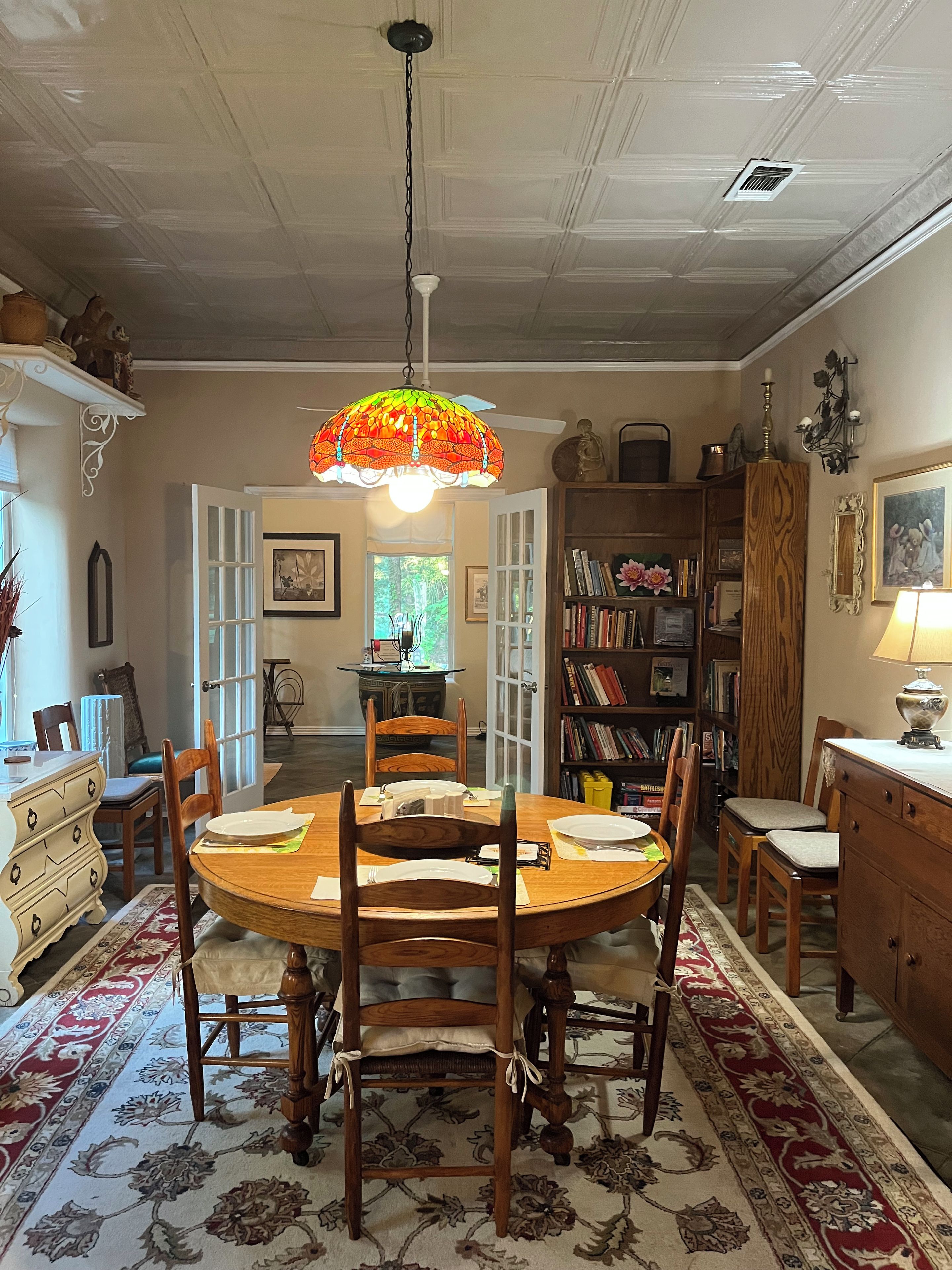 Ein Esszimmer mit einem runden Holztisch, Stühlen, einem gemusterten Teppich und einer farbenfrohen Glaskugel-Pendelleuchte.