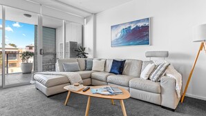 A modern living room with a sectional sofa, a coffee table, and a large window with blinds. - Kiama Coast | Beach, Balcony & Barbecue (Kiama)