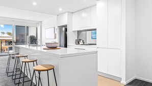 A modern kitchen with white cabinetry, a large island with stools, and a view of the outside. - Kiama Coast | Beach, Balcony & Barbecue (Kiama)