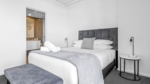 A modern bedroom with a large bed, a bedside table, a bench, and a view of a bathroom. - Kiama Coast | Beach, Balcony & Barbecue (Kiama)
