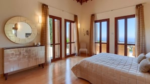 A bedroom with a large bed, wooden flooring, a round mirror, and a view of the sea through large windows. - Elegant Villa Retreat with Infinity Pool & Scenic Views (Paphos)