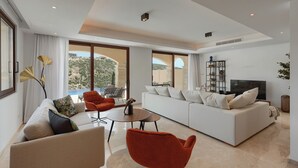 A spacious living room with a white sofa, red armchair, and a TV. Large windows offer a view of the outdoors.