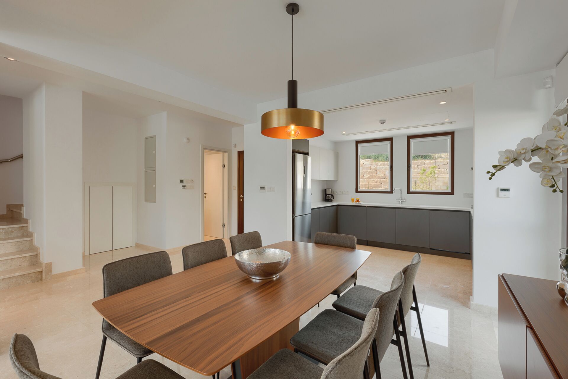 A modern dining area with a wooden table and chairs, a hanging pendant light, and a view of the kitchen and outdoor patio.