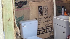 Une petite salle de bain rustique avec des toilettes blanches, un plafond en bois et des murs recouverts de sacs de jute sur lesquels figurent des publicités pour le café.