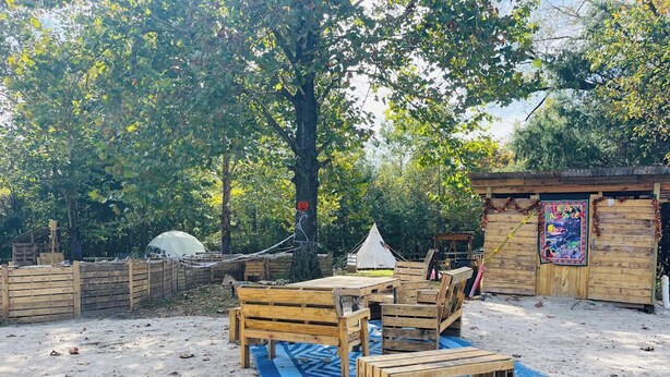 Un espace extérieur sablonneux aménagé avec du mobilier en bois, une tente et une structure en bois ornée d’une fresque colorée.