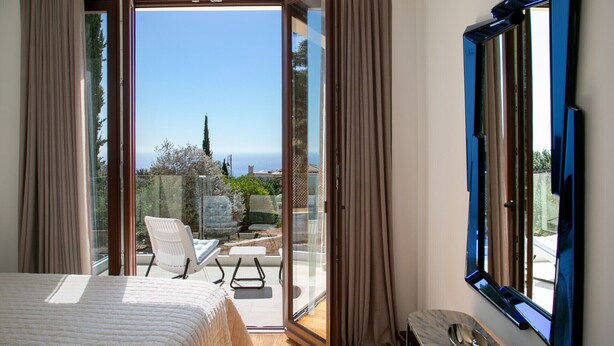 A bedroom with a bed, a seating area with chairs, a TV, and a view of the sea. - Spectacular Sea Views Await, Paphos Villa 1481 (Paphos)