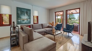 A modern living room with a sectional sofa, a glass coffee table, and two armchairs. There is a large painting on the wall, a small table with a vase, and a view of the outdoors through large windows.