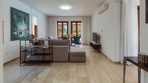 A modern living room with a sofa, a glass coffee table, and a TV. There are large windows with curtains, a ceiling light, and an air conditioning unit.