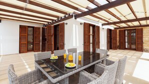 Un vaste espace extérieur aménagé pour les repas, avec une table en verre entourée de chaises en osier, de volets en bois et d’un sol carrelé.