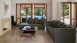 A modern living room with a grey sofa, a wooden coffee table, and a fireplace. There is a large window with a view of a swimming pool and outdoor furniture. - Unwind in Luxury: Your Perfect Family Escape (Paphos)