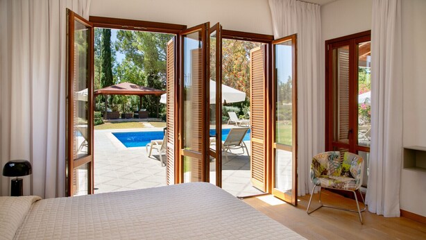 A bedroom with a large bed, a view of an outdoor swimming pool, and a chair by the window. - Unwind in Luxury: Your Perfect Family Escape (Paphos)