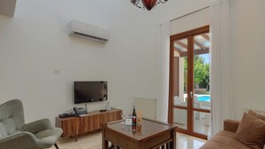 Ein modernes Wohnzimmer mit Flachbildfernseher, Couchtisch aus Holz, Sofa und Sessel. Eine Glastür öffnet sich zu einem Außenbereich mit einem Pool.