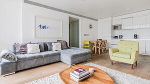Una sala de estar moderna con un sofá seccional gris, una mesa de centro de madera y un sillón amarillo. Se aprecia en el fondo un comedor con mesa y sillas de madera.