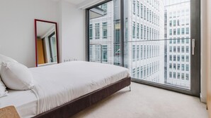 Un dormitorio moderno con una cama grande, un espejo y vistas de edificios altos a través de un ventanal de pared a techo.