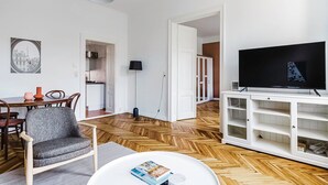 Een moderne woonkamer met een houten visgraatvloer, een flatscreen televisie op een witte standaard, een eethoek met een houten tafel en stoelen, en een keuken die zichtbaar is door een open deur.