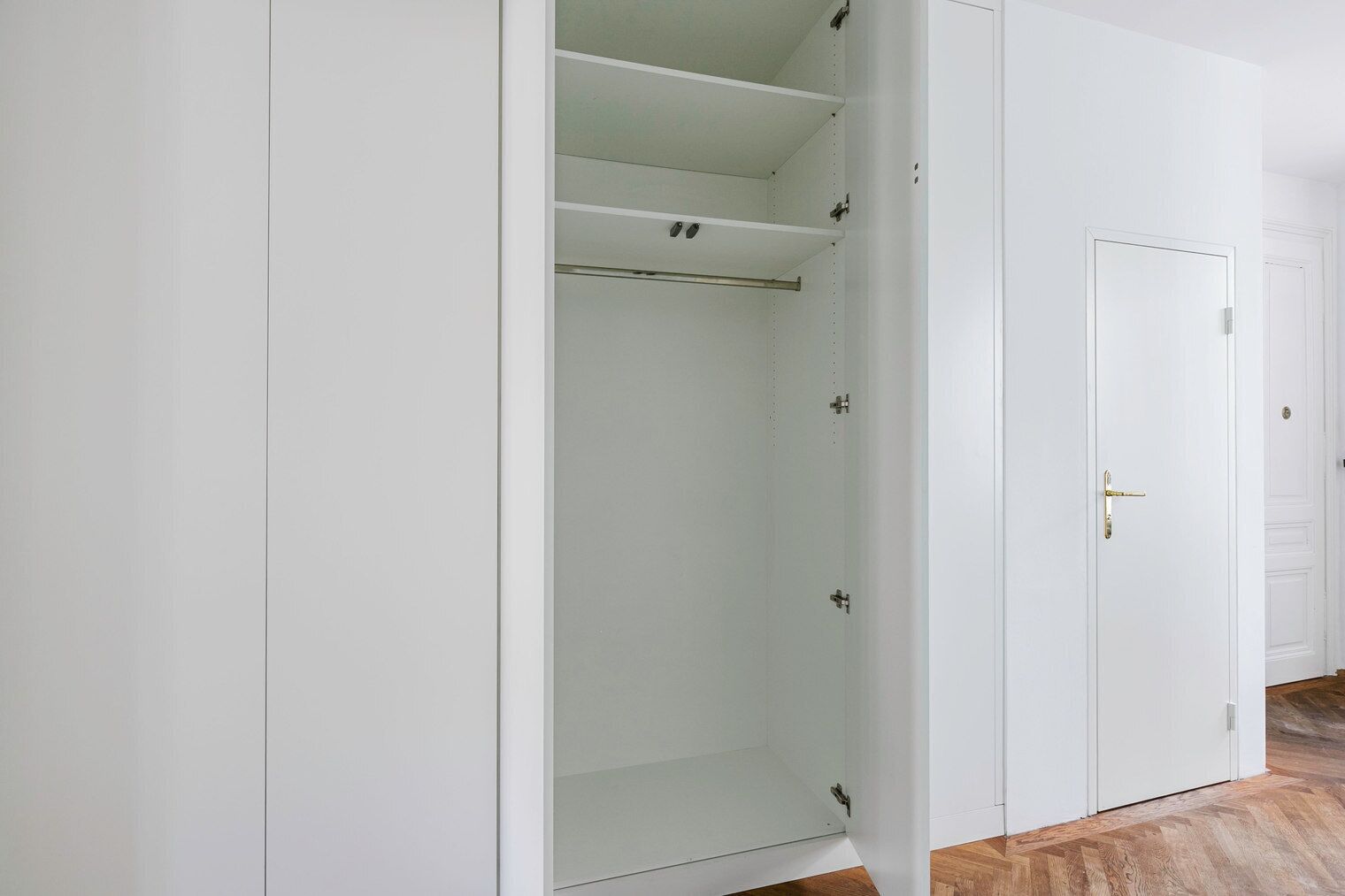 Een witte inbouwkast met meerdere planken en een kledingroede, een gesloten deur met een gouden handgreep, en een houten vloer.