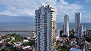A high-rise building with multiple balconies, surrounded by other tall buildings and a coastal view.
