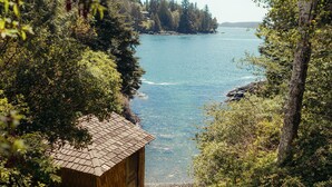 A wooden cabin with a sloped roof, surrounded by dense greenery and overlooking a calm body of water. - Maison Tofino - Private Oceanfront (Tofino)
