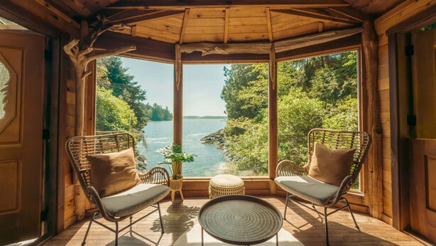 A wooden cabin with a large window overlooking a lake, furnished with wicker chairs and a small round table. - Maison Tofino - Private Oceanfront (Tofino)